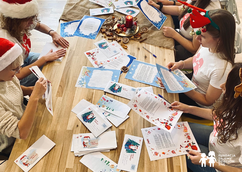 Grafika przedstawiająca młode kobiety siedzące przy stole. Kobiety pakują świąteczne listy do kopert. W prawym, dolnym rogu logo Fundacji Dajemy Dzieciom Siłę.